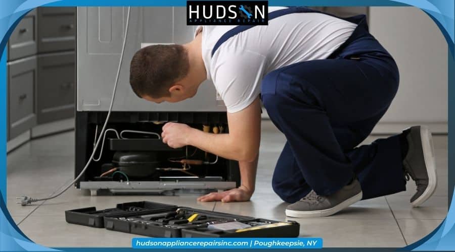 Technician crouching to inspect the back of an unplugged refrigerator with Hudson Appliance Repair branding.