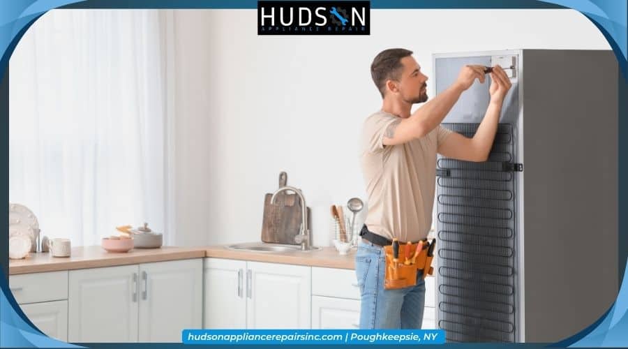 fridge-stopped-working - An appliance repairman wearing a tool belt uses a screwdriver to fix the back of a refrigerator in a kitchen.