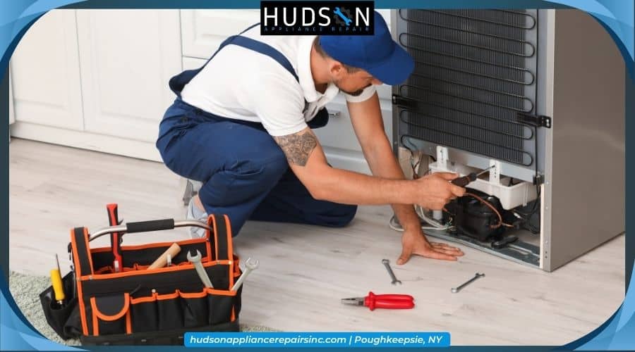 refrigerator-troubleshooting - An appliance technician kneeling on a kitchen floor, using a screwdriver to repair the lower back section of a refrigerator.